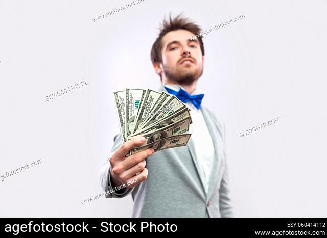 Take it! Portrait of confident attractive young businessman in gray coat and blue bow tie standing, proudly with head up and giving you many dollars