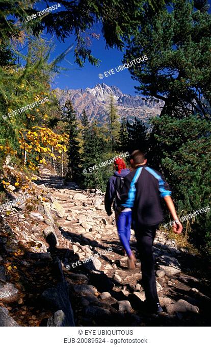 Trekkers on the Magistrala trail near Popradske Pleso. Rocky path lined by trees in Autumn colours with mountain peaks beyond