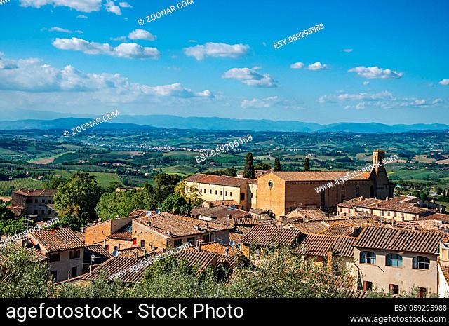 Overview of rooftops and church with green hills and blue sky at San Gimignano. An amazing medieval town famous for having several towers in its historical...