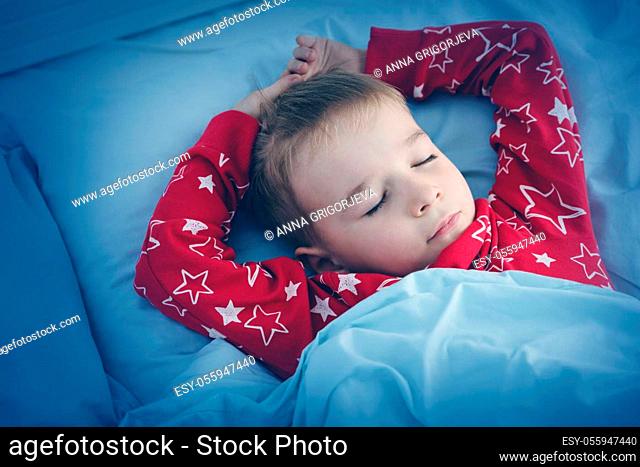 Sleepy boy lying in bed with blue beddings. Tired child in bedroom sleeping. Little kid lying asleep in red pajamas