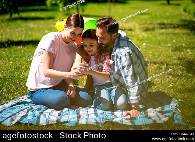 Father Mother And Daughter Looking At Photos On Smartphone In Summer Park. Sitting On Grass. Happy Family Concept