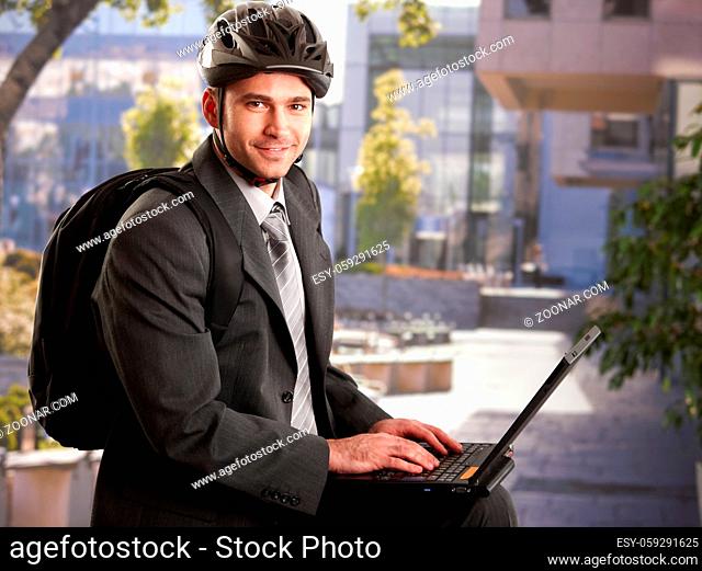 Portrait of young businessman wearing bike helmet, sitting in front of office building, using laptop computer