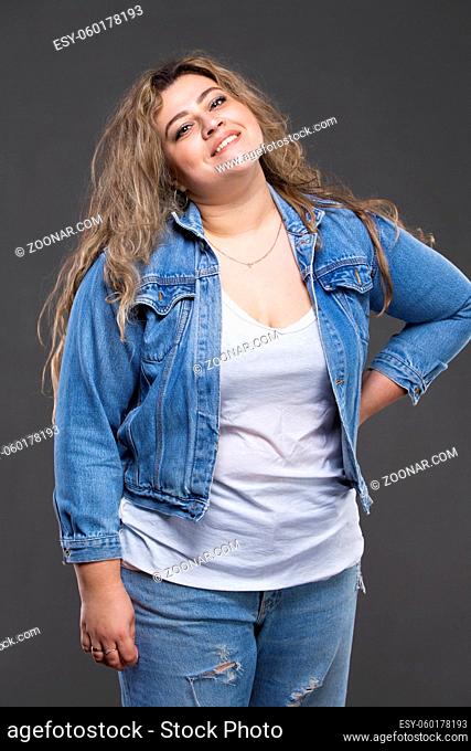 A beautiful fat woman in a denim jacket looks at the camera and smiles on a gray background