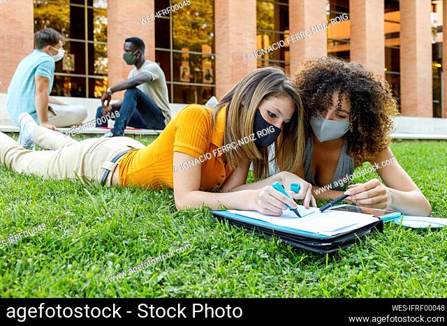Male and female friends wearing safety mask while studying over grass in university campus