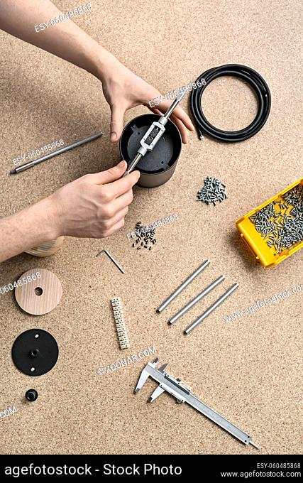 Man is using a t-wrench on a black metal cylinder on the light table. On the table there are billets, cable, plastic box with screws, tools, caliper, bushings