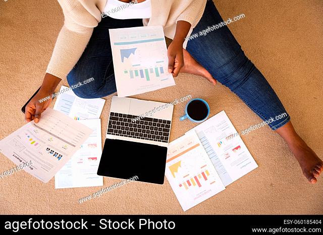 Happy african american woman working in bedroom, sitting floor using laptop and holding paperwork