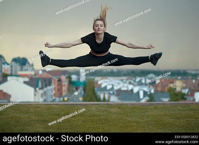 Young caucasian girl performs twine jumping over background cityscape, horizontal