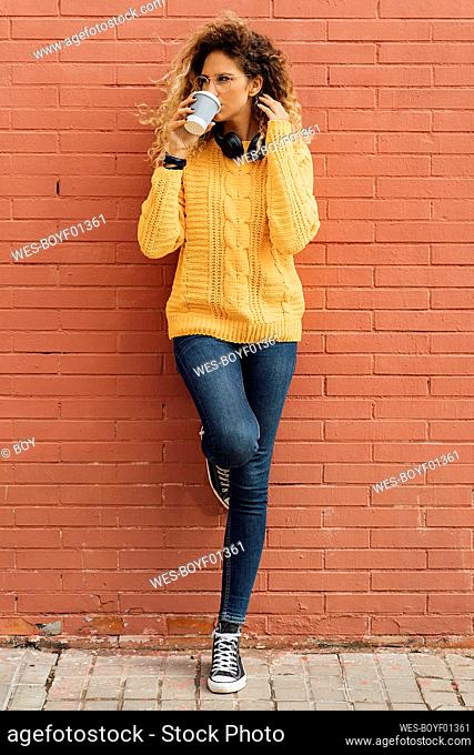 Beautiful young woman looking away while drinking coffee from disposable cup against red brick wall