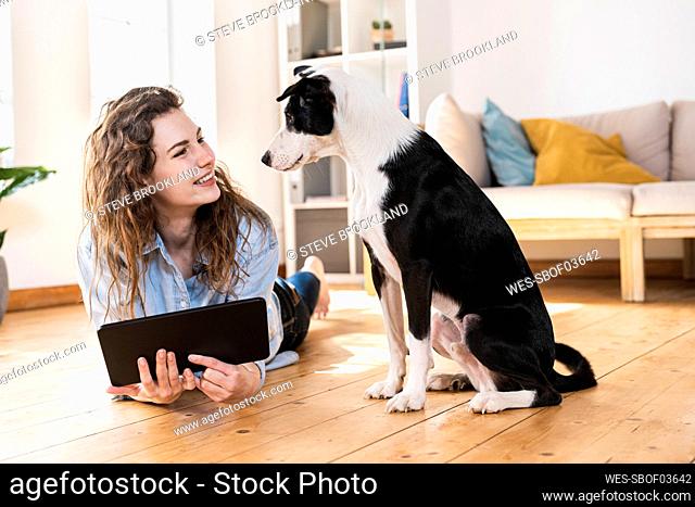 Happy woman with digital tablet looking at dog while lying on floor in living room