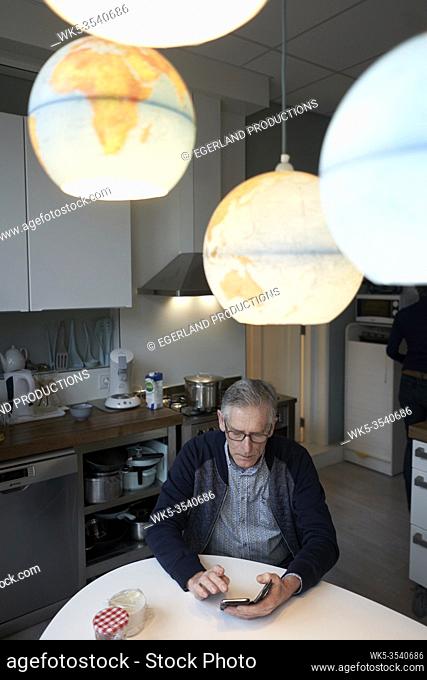 senior man using smartphone at breakfast table in kitchen under world globes