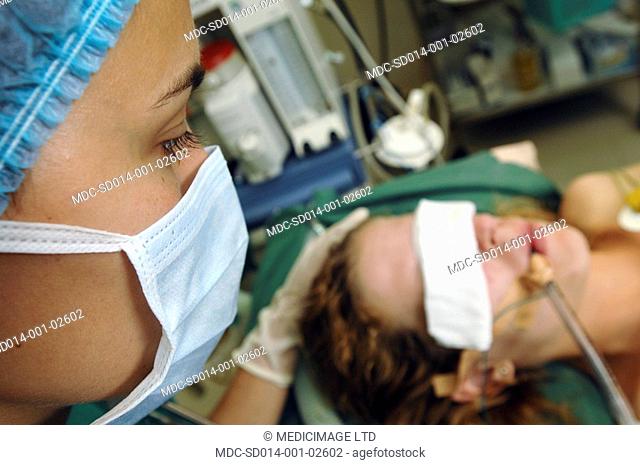 A female patient with her eYes covered at the start of a tracheotomy operation. As a surgeon is about to make an incision in her neck with a scalpel she...