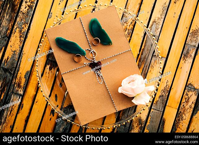 Beautiful brown envelope tied with a rope with rings, a rose and petals lies on a transparent tray on a wooden table. High quality photo