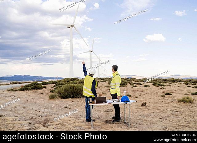 Engineer pointing at wind turbine standing with colleague at wind farm