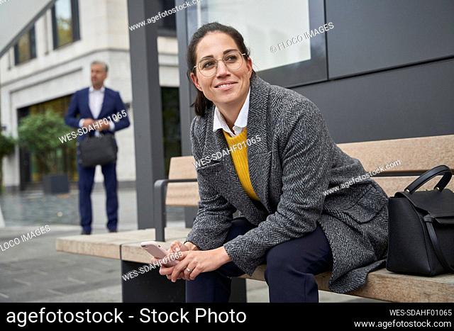 Smiling businesswoman sitting on bench waiting at bus stop while businessman standing in background
