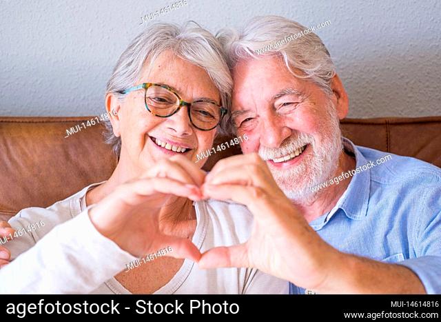 Portrait of cheerful senior couple embracing while doing heart shape gesture with hands. Elderly happy couple relaxing and posing in front of camera sitting in...