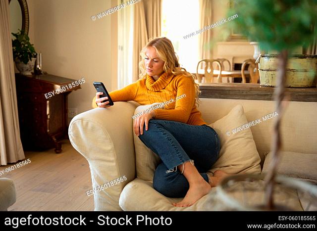 Caucasian woman sitting on couch relaxing in luxury living room using smartphone