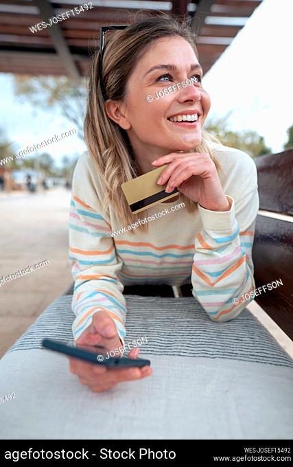 Thoughtful young woman lying on bench holding credit card and smart phone
