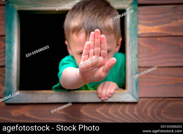 Little Caucasian boy looking through the window of a wooden toy house in a outdoor playground and showing stop hand sign