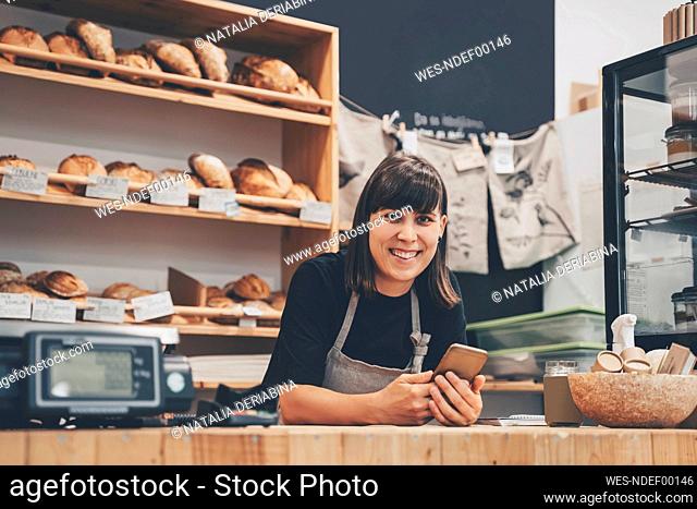 Smiling store owner with mobile phone leaning on checkout counter in shop