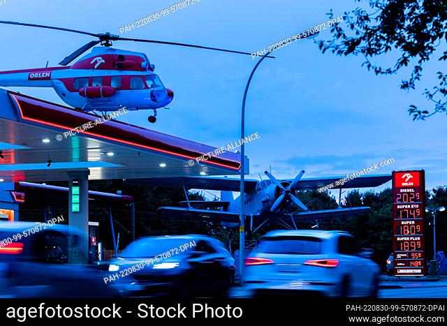 30 August 2022, North Rhine-Westphalia, Leverkusen: Cars refuel and drive past a gas station with an old helicopter on its roof and a historic Antonov AN2...