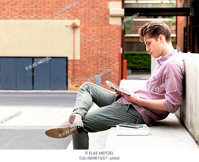 Young man sitting on step, outdoors, reading book