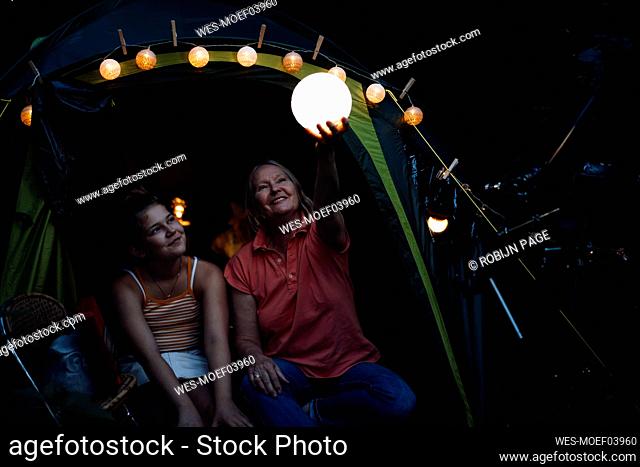 Smiling woman holding illuminated lantern while sitting with girl at backyard
