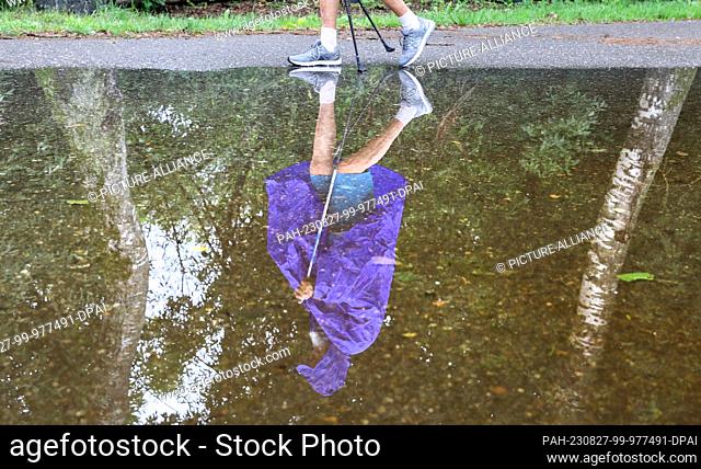 27 August 2023, Baden-Württemberg, Bad Buchau: A man in rain gear is reflected in a puddle. More precipitation is predicted for the next few days Photo: Thomas...