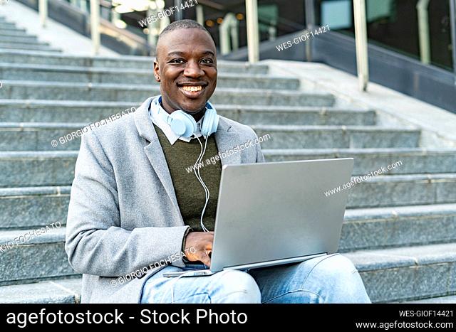 Smiling businessman with headphones and laptop sitting on steps