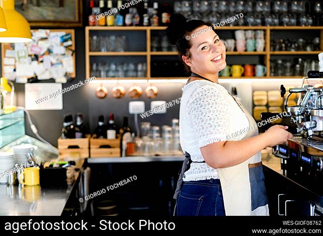 Smiling female barista standing by coffee maker while working in cafe