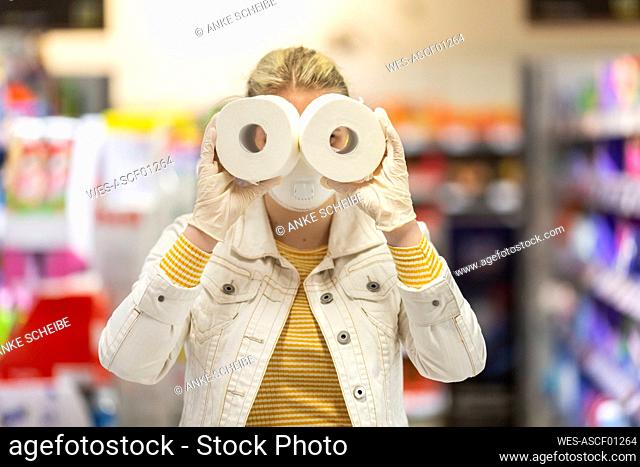 Teenage girl wearing protectice mask and gloves holding looking through holes of toilet rolls at supermarket