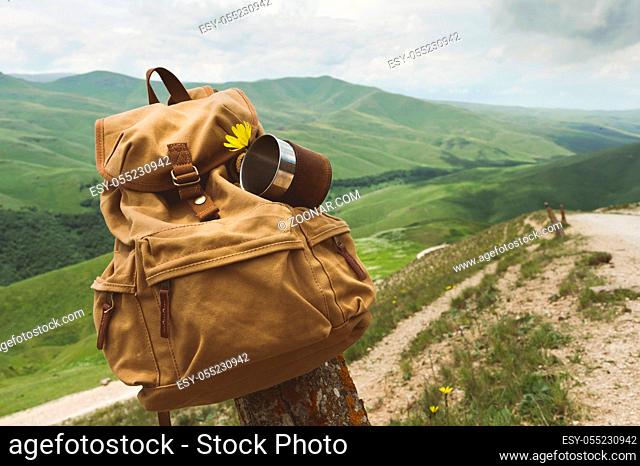 Hipster yellow vintage backpack with a mug fixed on it with a mug close-up front view. Traveler's travel bag in the background of a mountain landscape and a...