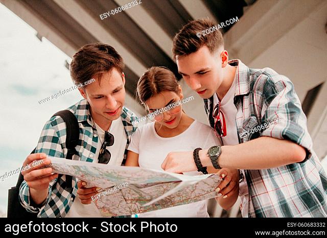 Team Of Teenagers Is Studying The Map Of The City. Two Cute Guys And A Girl With Backpacks Considering The Map And Choosing The Route Of Travel
