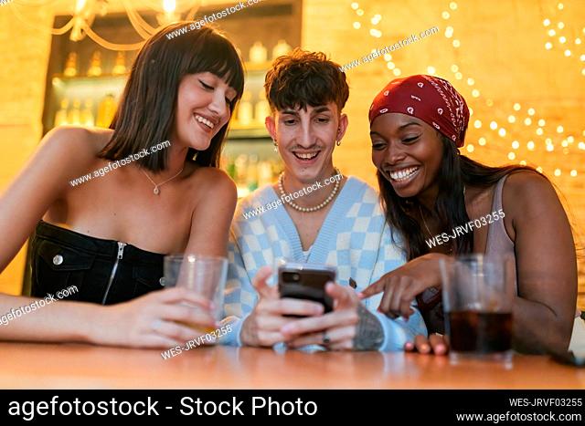 Happy young man sharing smart phone sitting amidst friends in restaurant