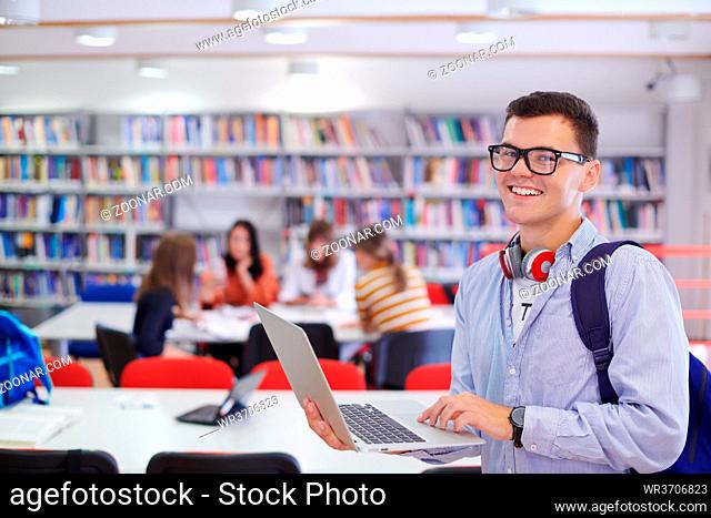 a young student with glasses stands in the school library with a smartphone, headphones and a laptop in his hand