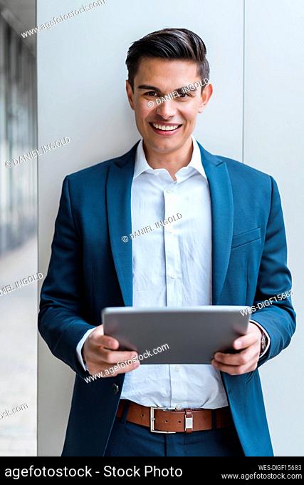 Smiling male professional holding digital tablet in front of wall at corridor