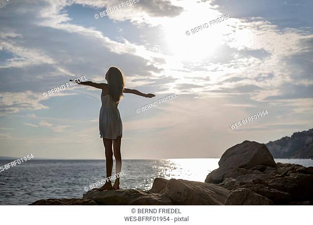 Croatia, Lokva Rogoznica, back view of girl with arms outstretched standing on a rock at dusk