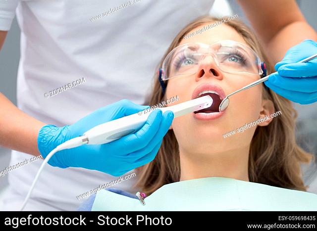 Pretty girl with opened mouth in patient bib and protective eyewear. Next to her there is a dentist in a white uniform with blue latex gloves