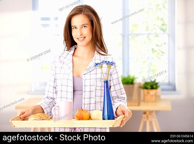 Happy woman standing with breakfast tray in living room, smiling at camera