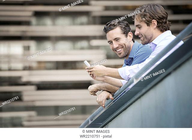Two men leaning on a railing, one holding a smart phone
