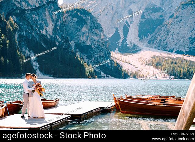 The bride and groom walk on a wooden boat dock at Lago di Braies in Italy. Wedding in Europe, at Braies lake. Newlyweds walk, kiss
