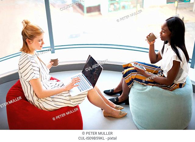 Two businesswomen drinking takeaway coffee sitting on beanbags looking at laptop and smartphone
