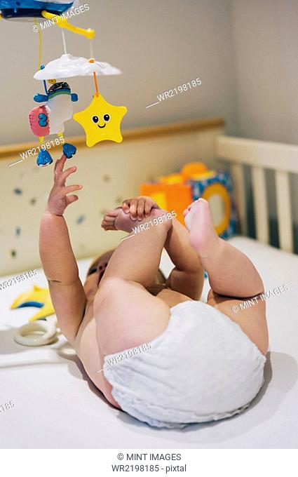 A baby girl in a diaper lying in a cot reaching up to a colourful mobile hanging above her