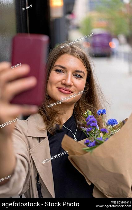 Smiling woman taking selfie with flower bouquet while standing on footpath