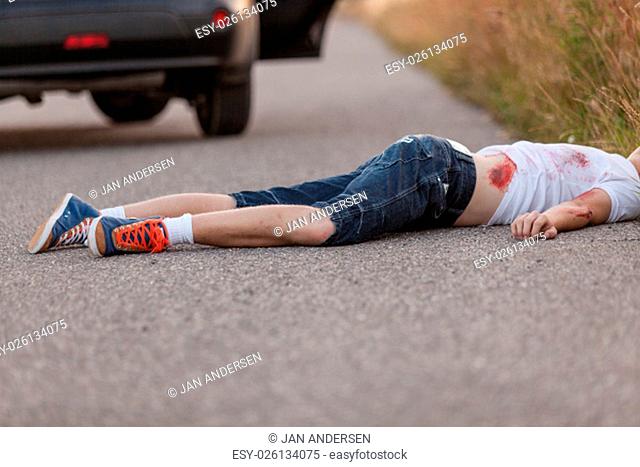 Young boy run over by a car lying face down in the road bleeding from his injuries with a car with an open door visible behind