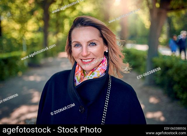 Attractive elegant middle-aged woman with a friendly smile standing in a tree-lined lane in the shade in a close up head and shoulders view