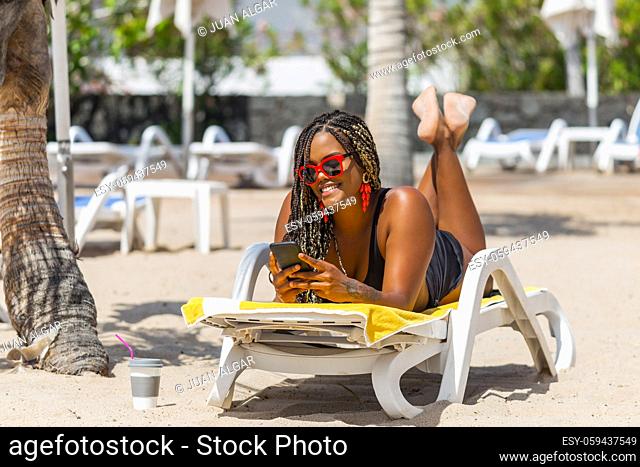 African American woman on the beach lying on a deck chair using phone. Cheerful adult woman enjoying a summer day at the beach
