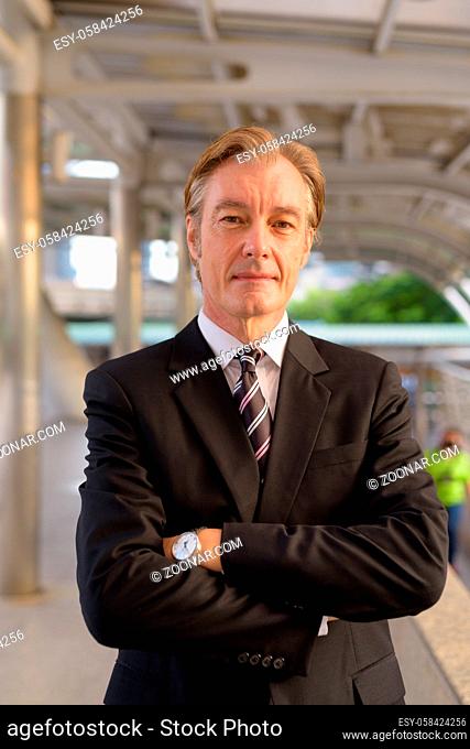 Portrait of mature handsome businessman in suit at skywalk bridge in the city outdoors