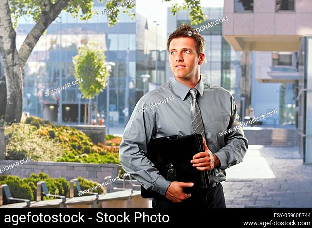Smiling businessman holding briefcase standing outside office building in sunshine