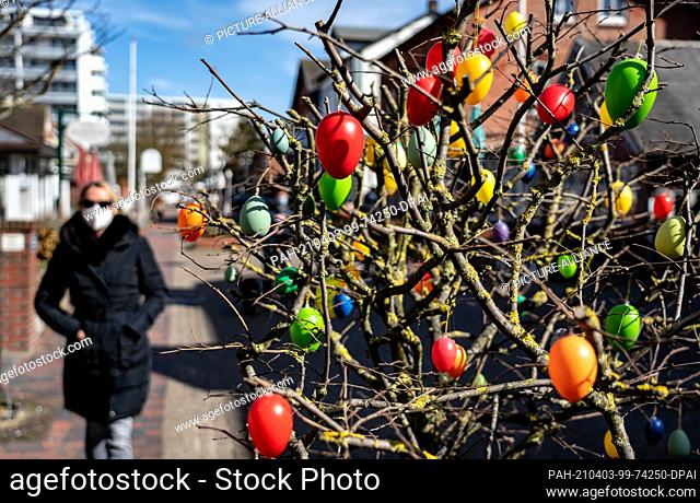 03 April 2021, Schleswig-Holstein, Westerland/Sylt: A woman with a mouth-nose protection walks past a bush with Easter eggs in the city centre of Westerland