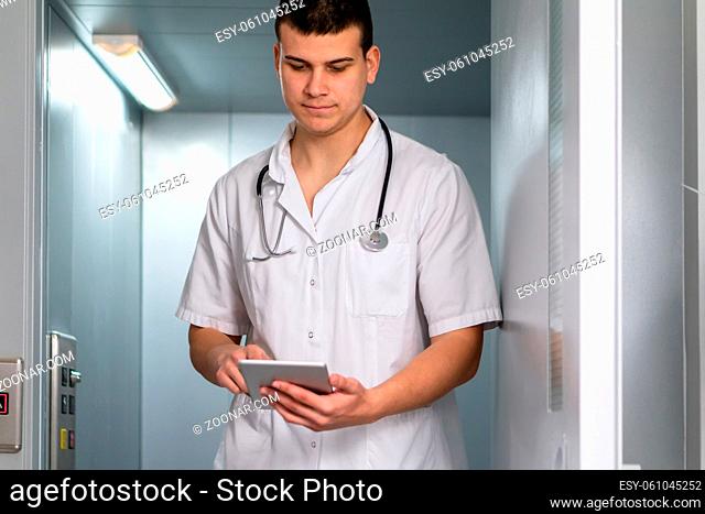 photo of male doctor in uniform with stethoscope coming out of the elevator and using computer tablet in hospital. Modern medical concept